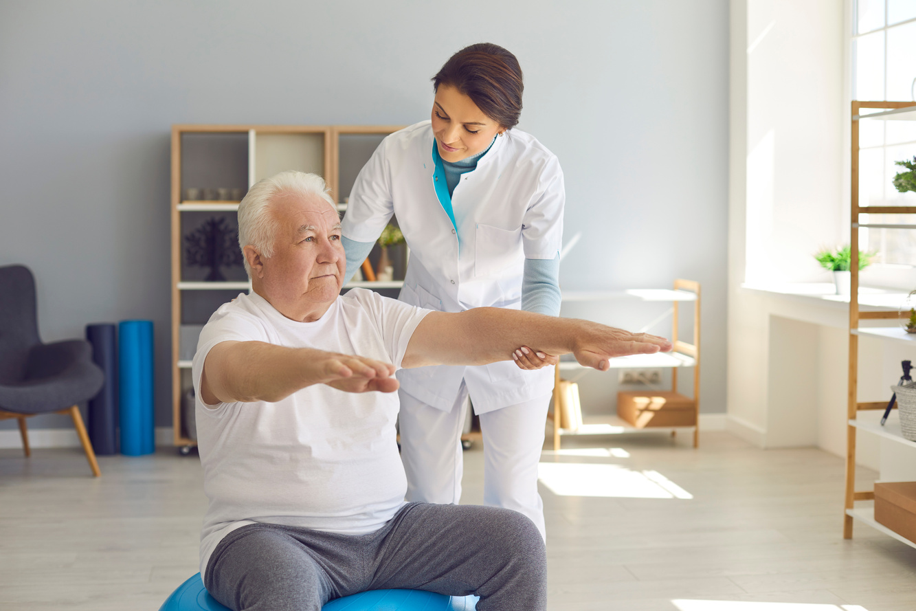 Female Physiotherapist Trains Senior Man in Room Supporting His Outstretched Arms.